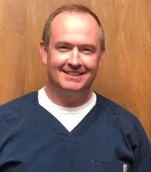 Male dental professional smiling at camera wearing navy scrubs against wooden wall background.