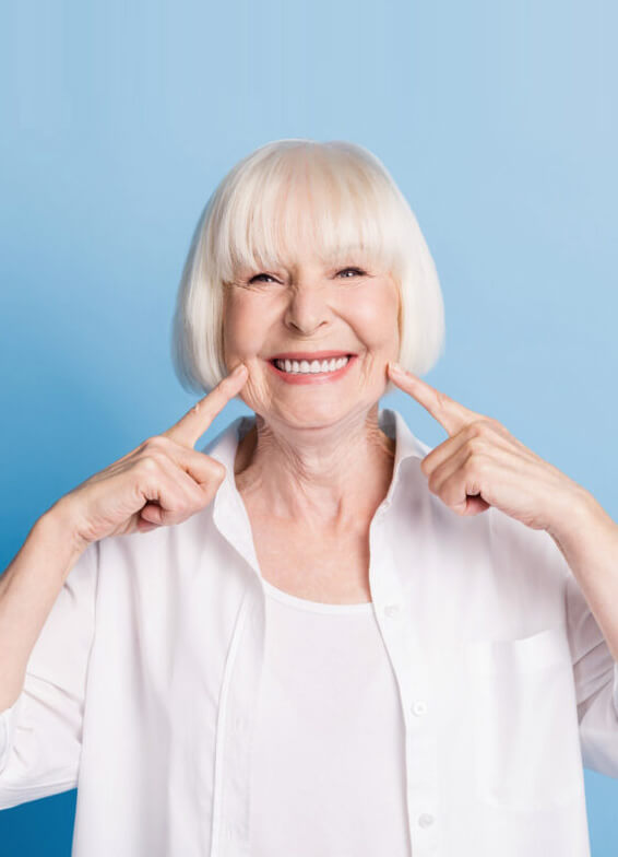Smiling mature woman pointing to her teeth against blue background, dental care portrait.