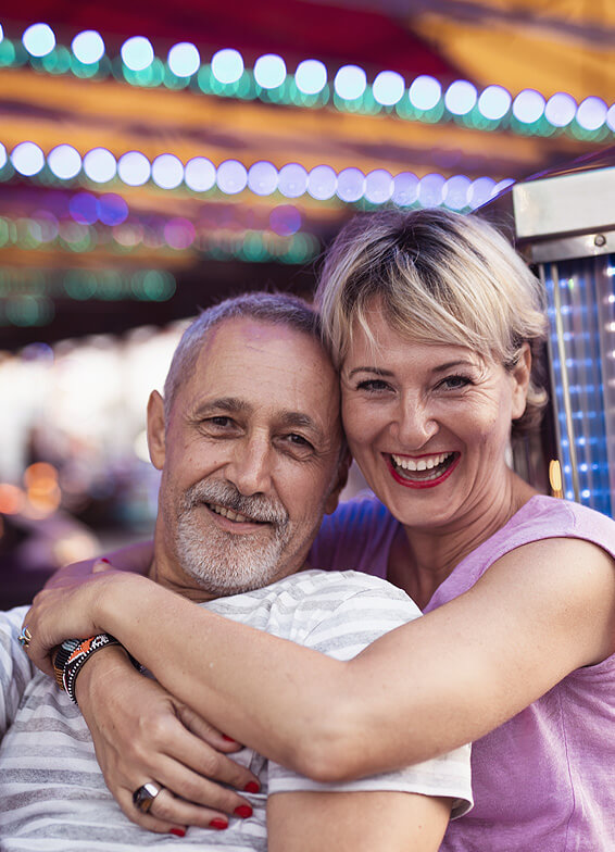 Smiling couple embracing at amusement park with colorful lights in background.