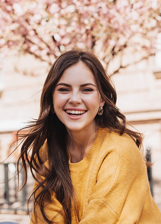 Woman in yellow shirt smiling outdoors with blurred pink flowers in background.
