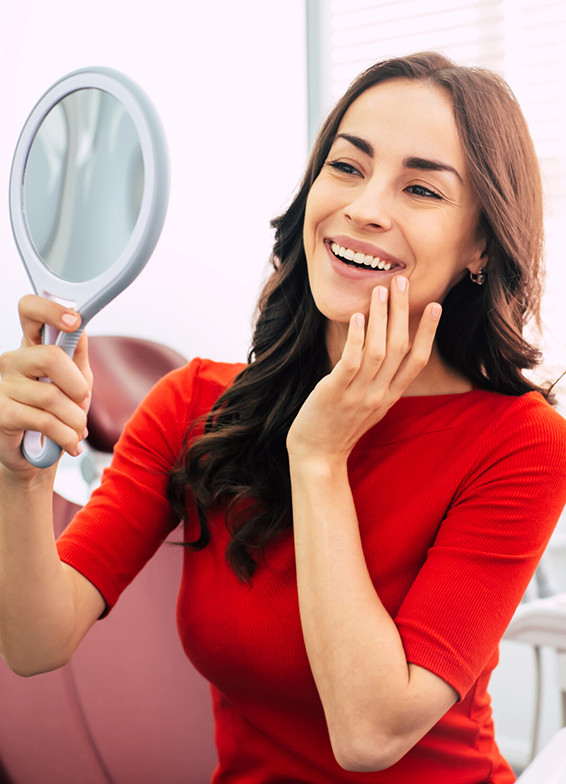 Woman in red shirt holding mirror, smiling at her reflection in dental office setting.
