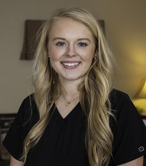 Professional headshot of smiling woman with blonde hair wearing black shirt in dental office setting.