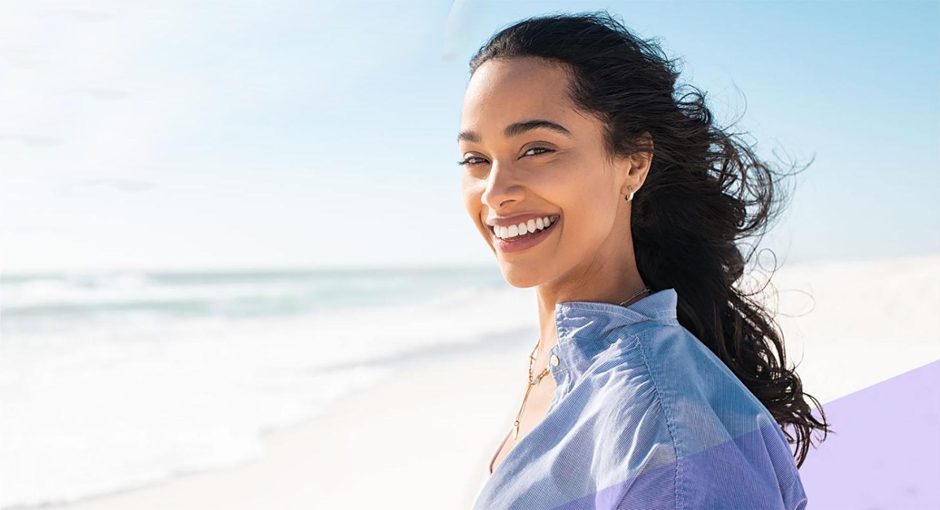 Woman smiling at beach wearing blue shirt, dental health and wellness lifestyle portrait.